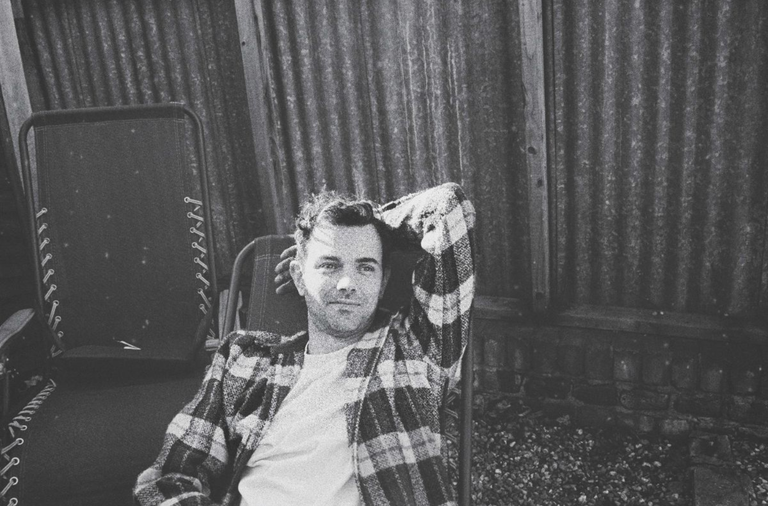 Black and white press photo of Tom Iansek of #1 Dads wearing a plaid jacket and reclining outdoors by a corrugated fence.